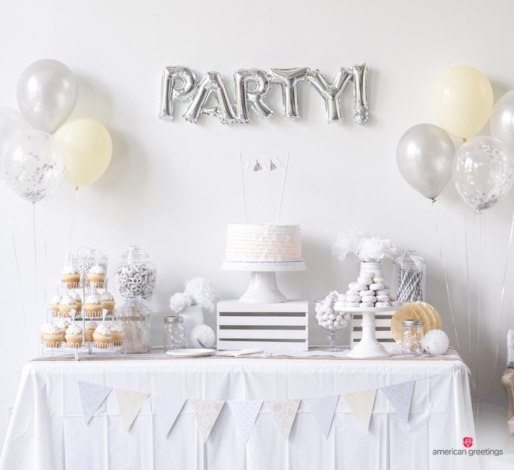 A picture of a white party table complete with white cupcakes, white paper flowers,donuts, and chocolate pretzels placed on a white tablecloth decorated with a white triangle banner and balloons.